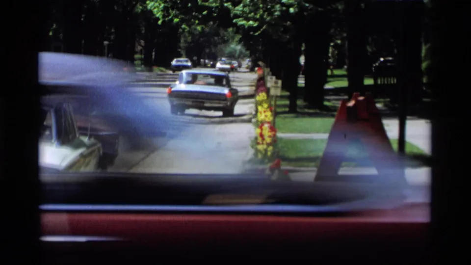 1969 Holland, Michigan frame looking through a vehicle window as cars travel along a shaded street near a sidewalk sign.