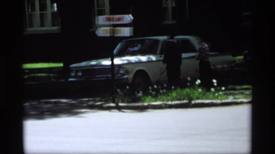 1969 Holland, Michigan home-movie frame showing a white sedan by a Tulip Lane sign and two people on a sunny residential street.