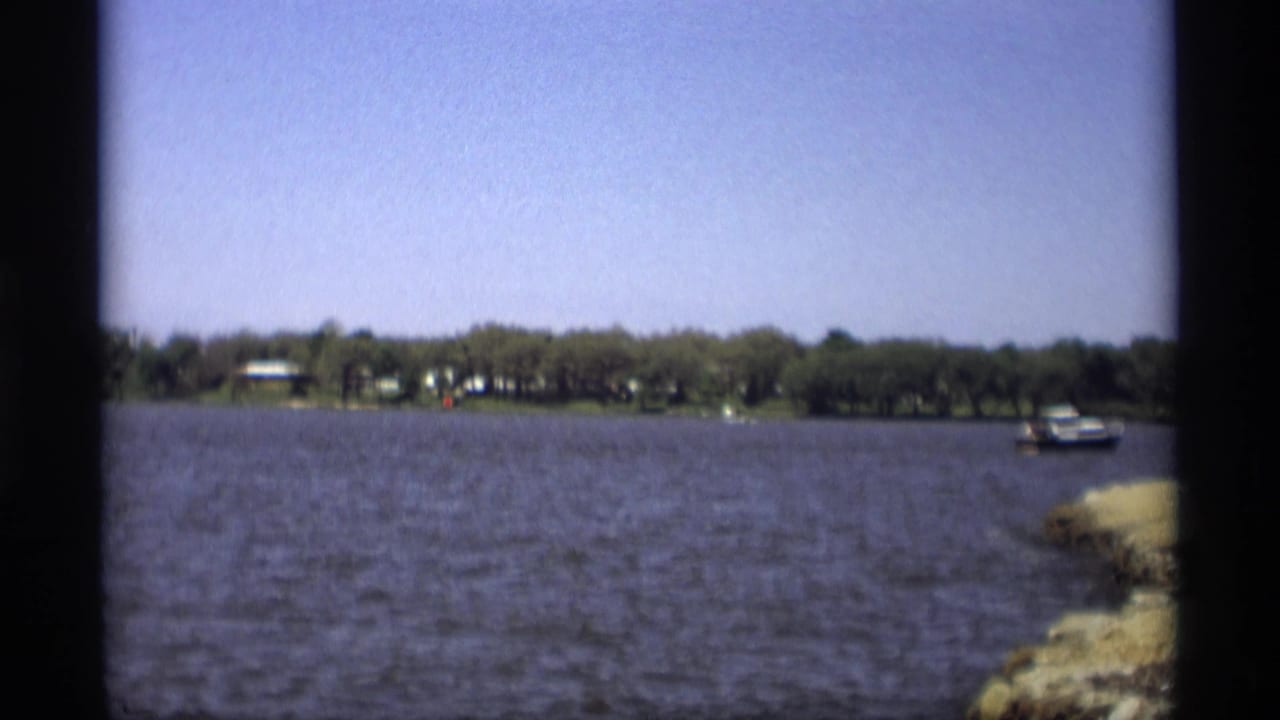 1969 8mm home-movie view of a Holland, Michigan lake with distant houses, trees, a small boat, and rocky shoreline.