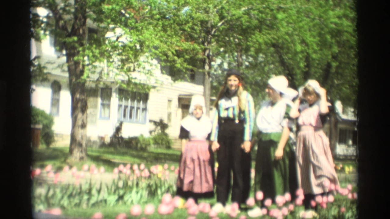 In Holland, Michigan in 1969, four people in Dutch-style costumes pose in a sunlit tulip garden with trees and a white house behind them.