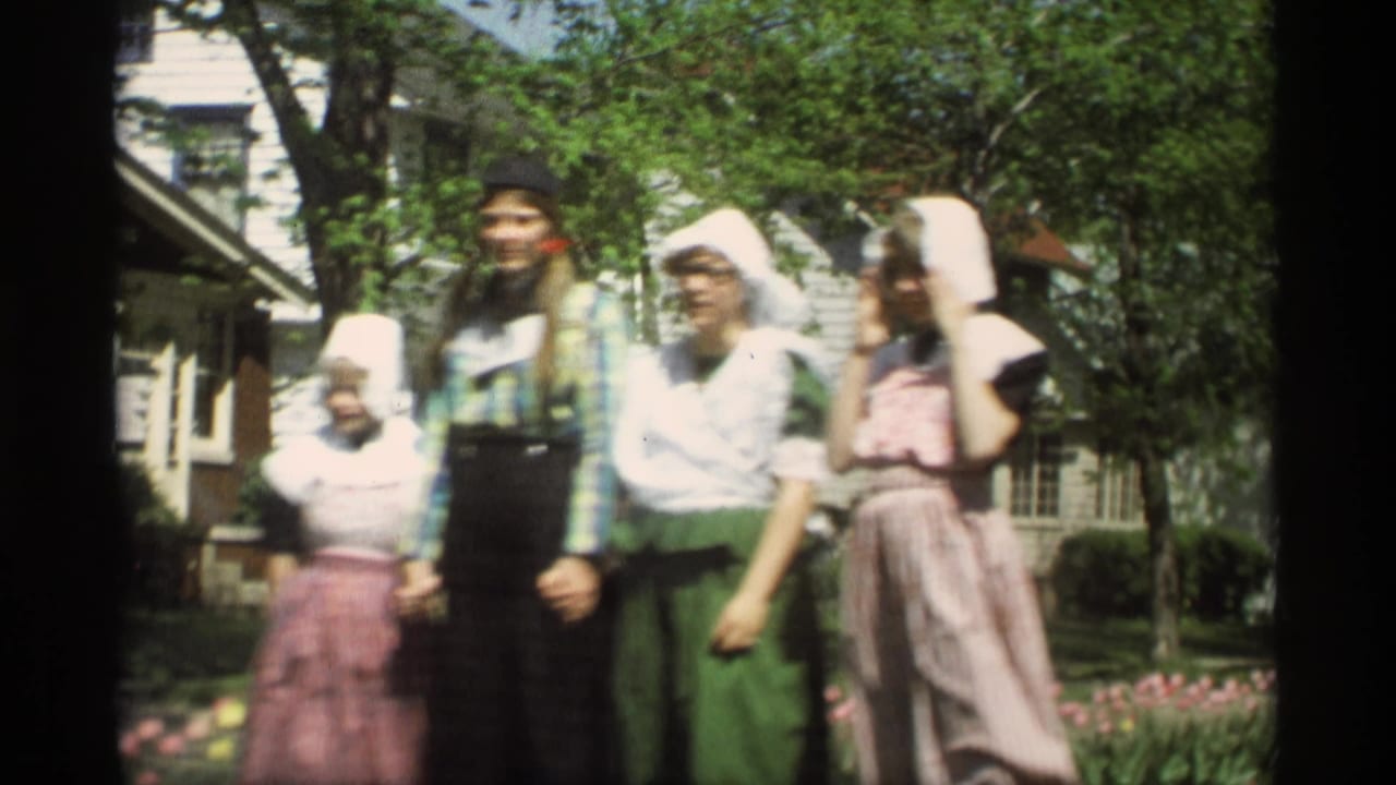 A closer 1969 Holland, Michigan home-movie view shows people in Dutch-style costumes standing by tulips under trees near houses.