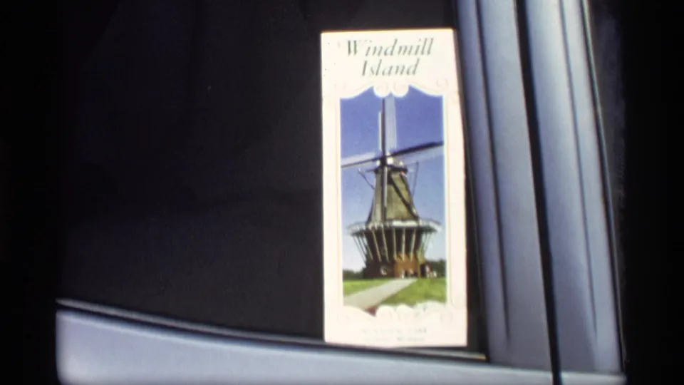 The camera holds on the Windmill Island brochure inside a car, with the windmill photo and printed title visible in soft 8mm focus.