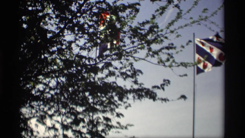1969 Holland, Michigan home-movie frame with a blue-and-white striped flag partly framed by foliage.