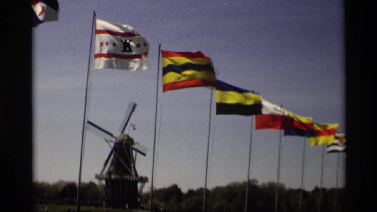 Colorful flags on tall poles wave near a Dutch-style windmill under a clear sky.