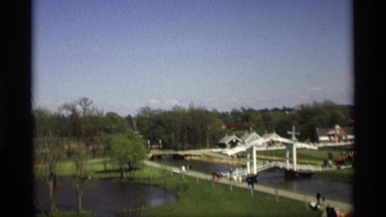 Elevated park view with a white drawbridge, river, lawns, trees, paths, and distant people.