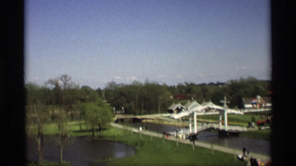 1969 8mm view in Holland, Michigan, showing a white drawbridge over water, green lawns, trees, small buildings, and distant park visitors.