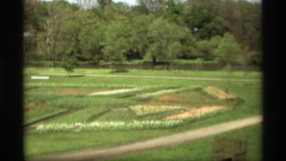 1969 Holland, Michigan park frame with a decorative flower bed, curved path, green lawn, pond, and trees in soft 8mm color.