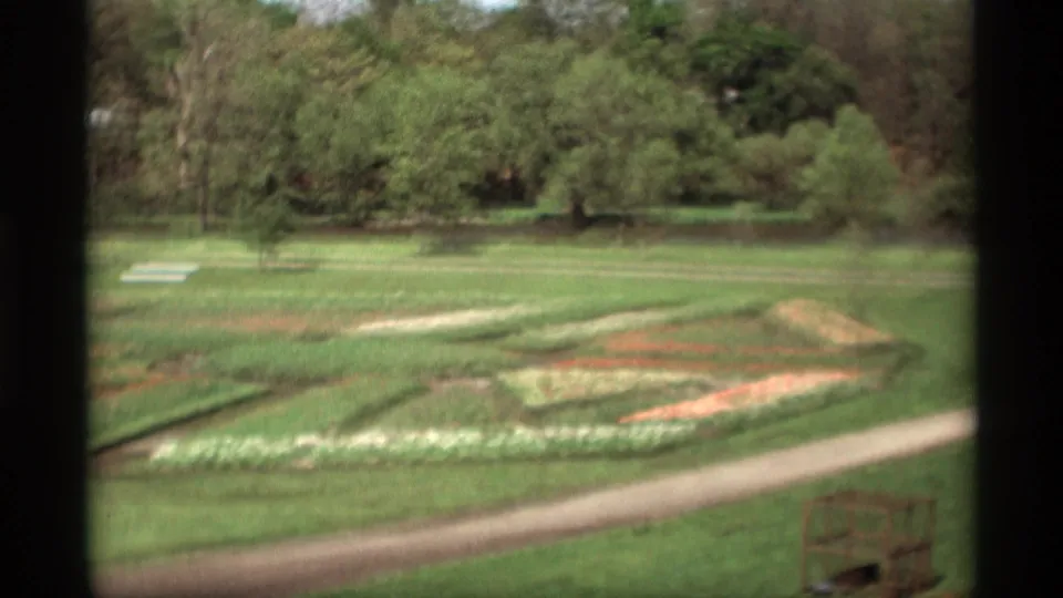 Vintage 8mm garden view in Holland, Michigan, with patterned flower beds, a pale path, and trees in the background.