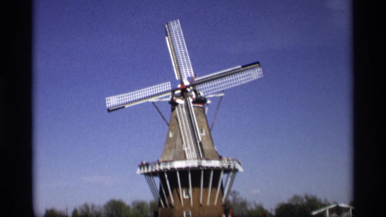 Dutch-style windmill with white sails against a blue sky and tree line.
