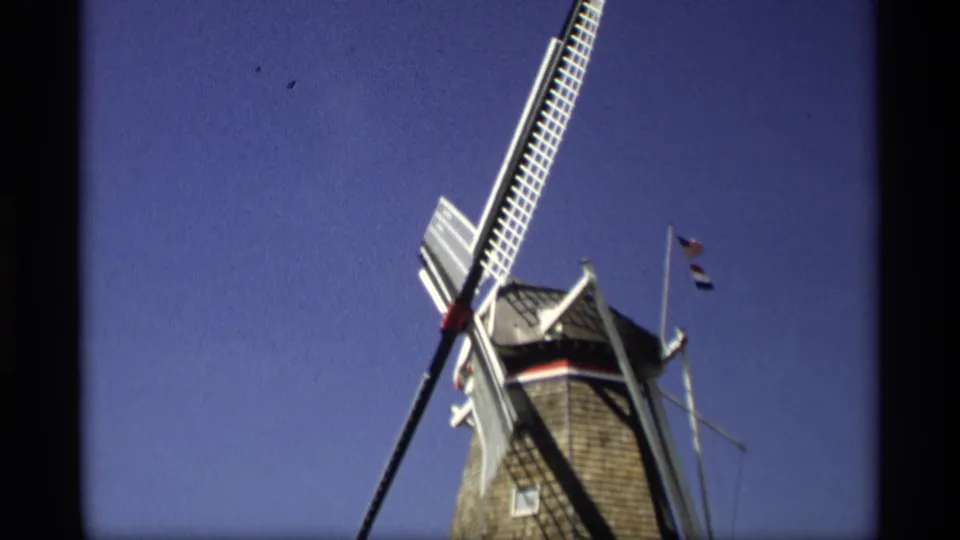 1969 Holland, Michigan frame showing the upper windmill, angled blades, flagpoles, and small U.S. and Dutch flags in clear daylight.
