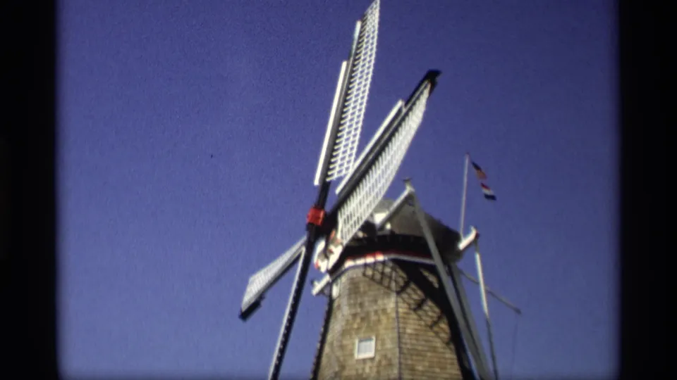 Low-angle 1969 8mm frame from Holland, Michigan, with the windmill tower, white blades, and flags visible against blue sky.
