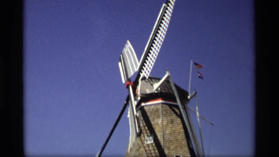 Final 1969 Holland, Michigan frame showing the windmill top, long white blade, dark support, and small flags in bright sun.