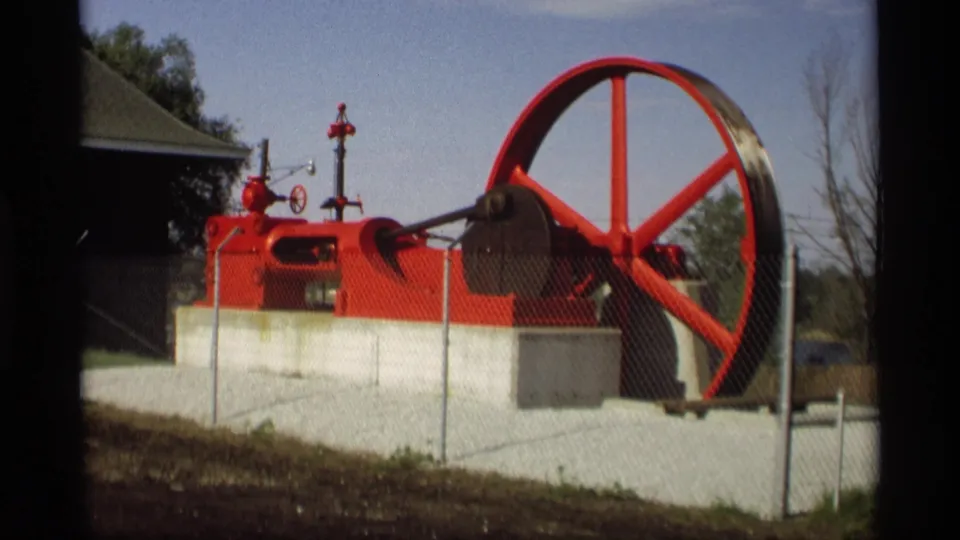 Holland, Michigan home-movie frame of a red engine or pump display with a large wheel and fence in the foreground.