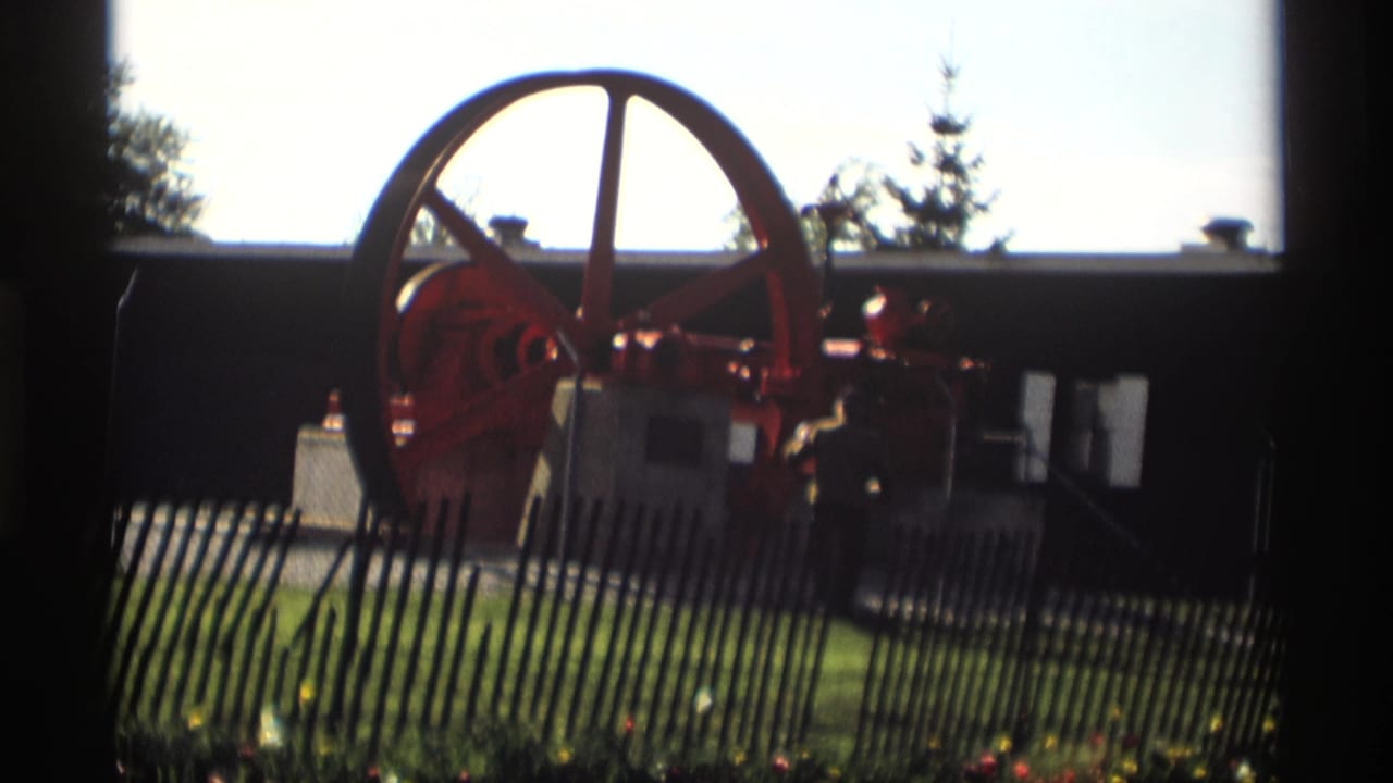 Red flywheel engine behind a dark picket fence on a sunny lawn.