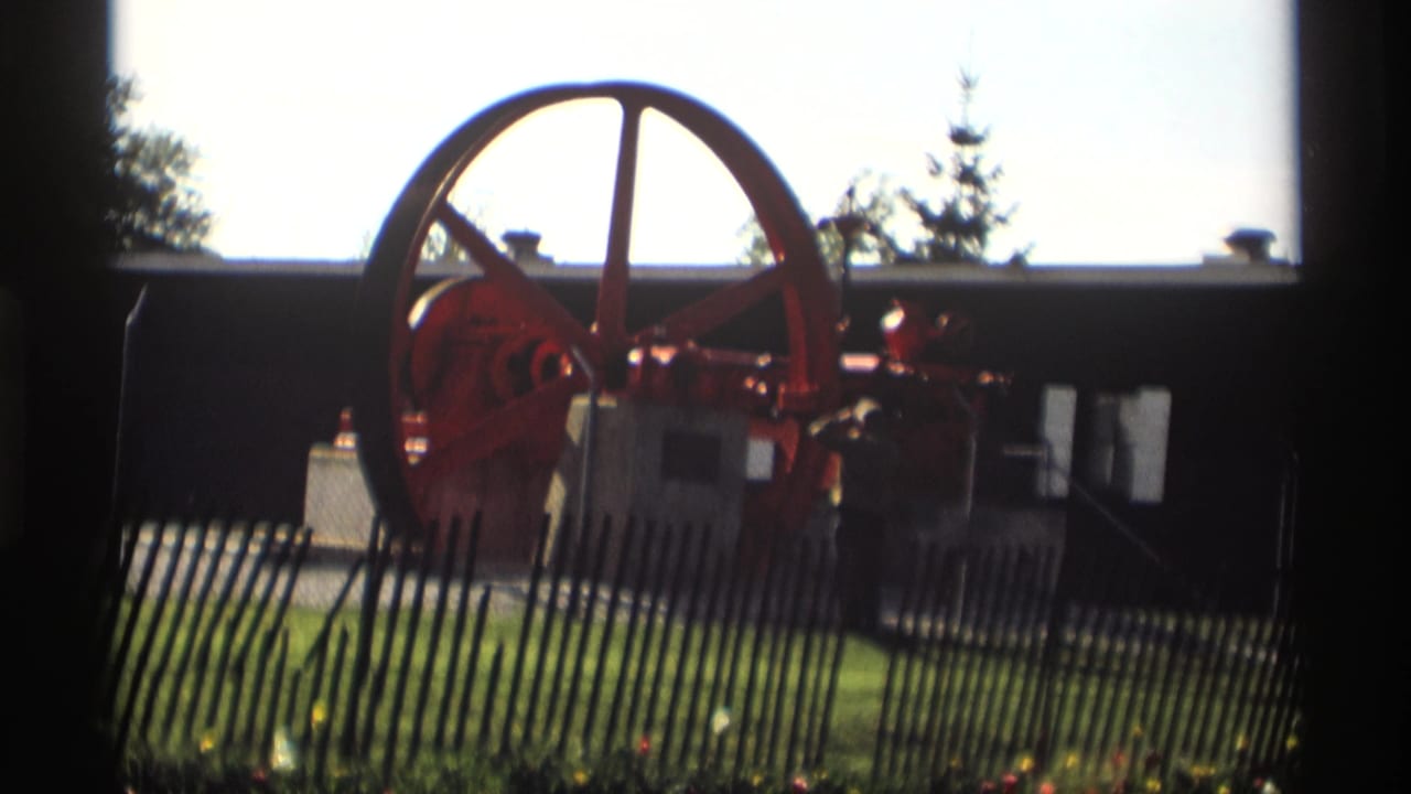 A man stands near the red flywheel engine in a 1969 Holland, Michigan outdoor home-movie scene.