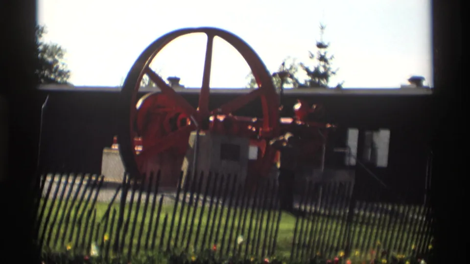 A man stands near the red flywheel engine in a 1969 Holland, Michigan outdoor home-movie scene.