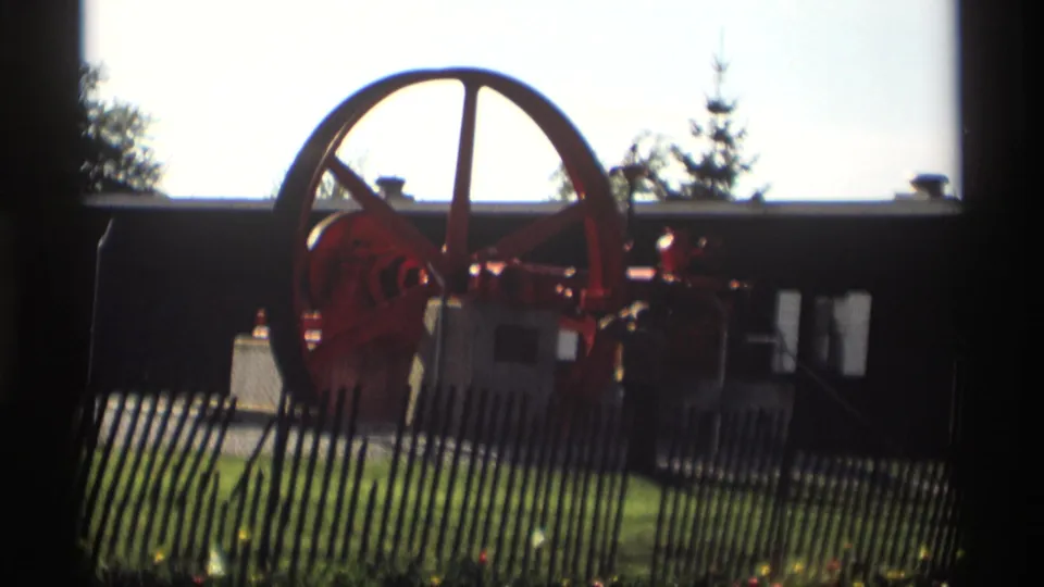 The 8mm frame holds on the red industrial engine behind fencing in Holland, Michigan, with lawn and trees beyond.