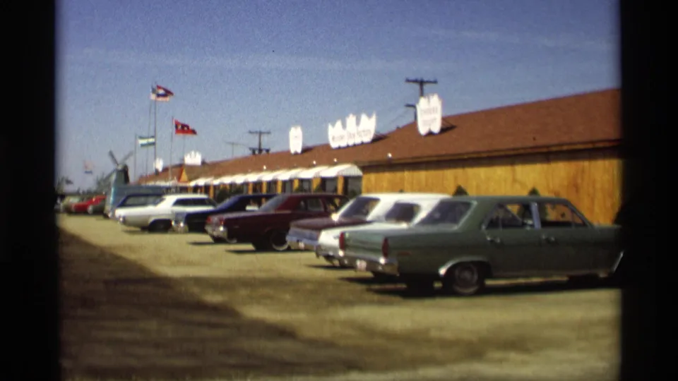 1969 Holland, Michigan roadside scene with a long red-roofed building, flagpoles, signs, and late-1960s cars in front.