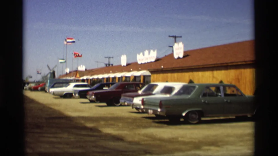 Home-movie view of a Holland, Michigan roadside business with flags, signage, and a row of 1960s cars in the lot.