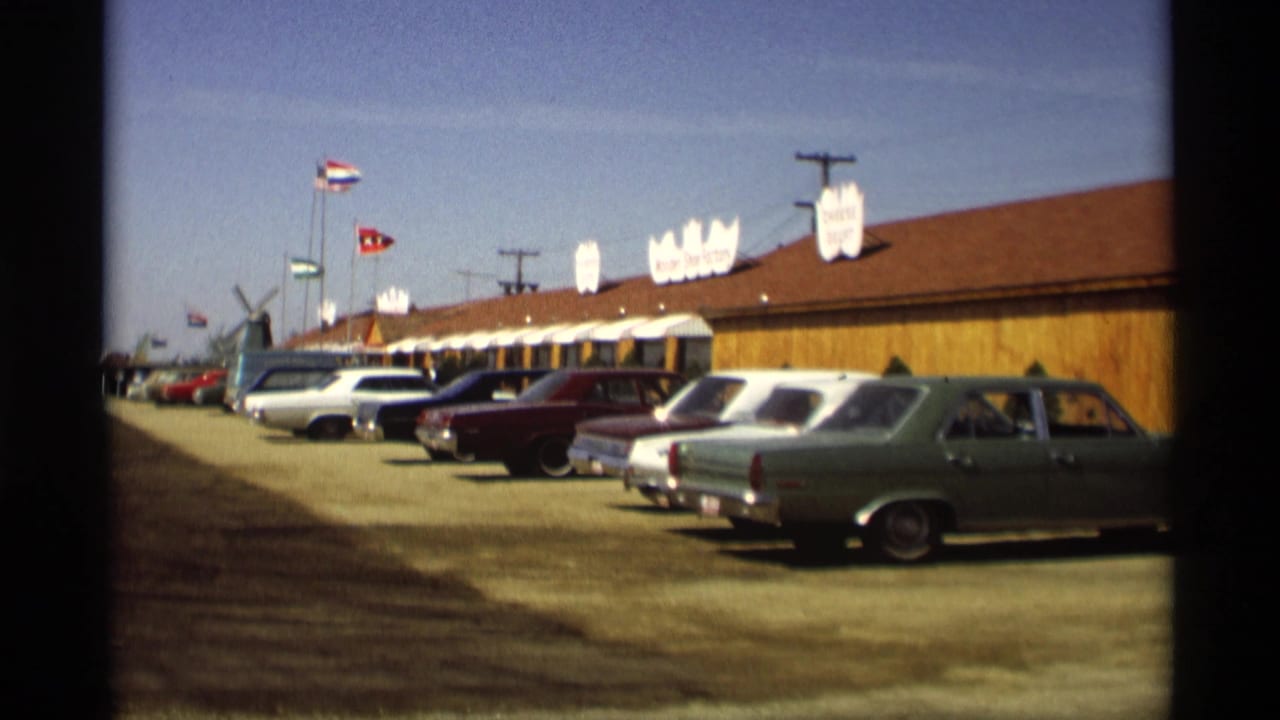 Rows of late-1960s cars parked beside a long wooden building with flags above it.