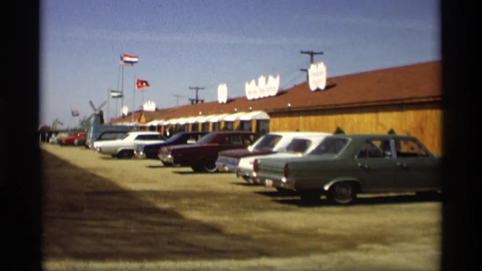 Late-1960s cars sit outside a long wooden building in Holland, Michigan, with flags and a distant Dutch-style windmill shape visible.