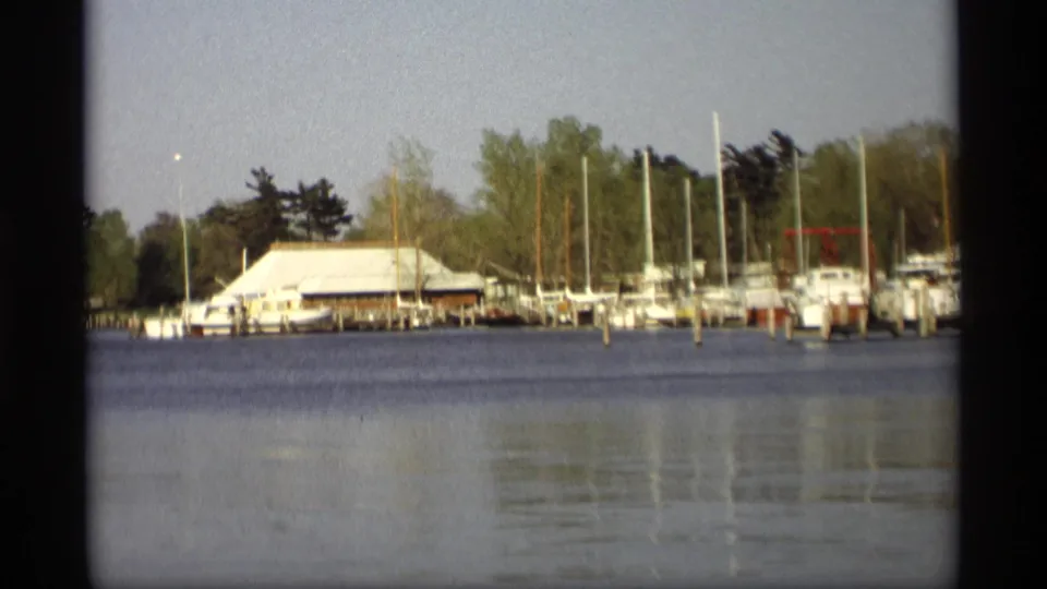 Holland, Michigan marina view from 1969 home-movie footage, showing docked boats, sailboat masts, shoreline trees, and reflective water.