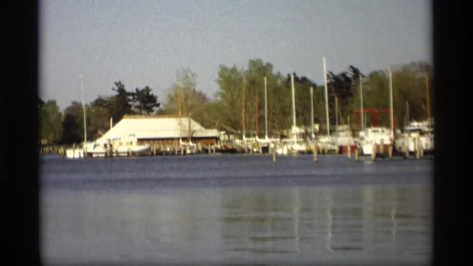A 1969 Holland, Michigan waterfront frame shows a small marina with moored boats, tall masts, trees, and calm foreground water.