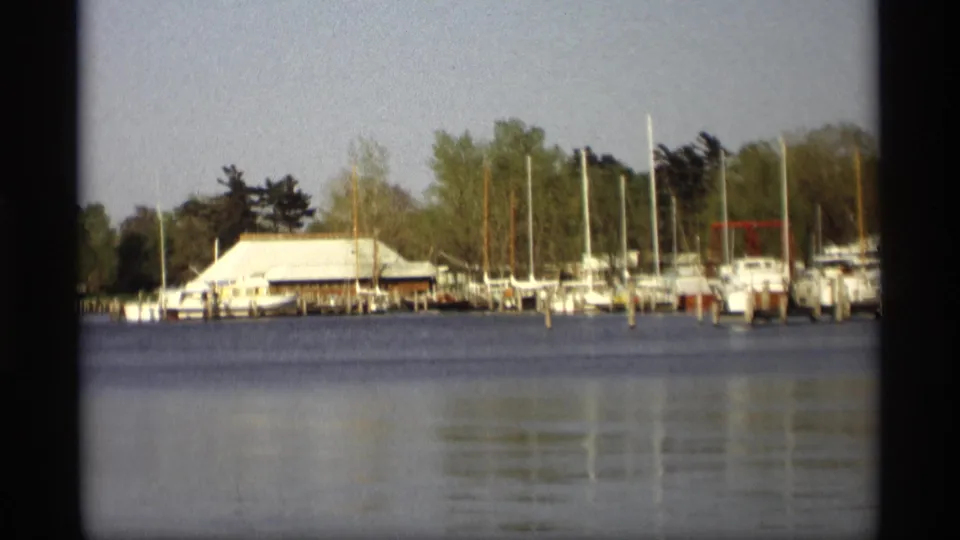 1969 Holland, Michigan 8mm frame showing docked white boats, tall masts, shoreline trees, and low marina buildings across calm water.