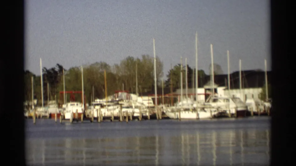 1969 Holland, Michigan marina view with docked white boats, vertical masts, shoreline trees, and muted 8mm color across still water.