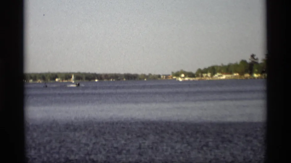 Distant sailboats appear on calm water in Holland, Michigan, with a wooded lakeshore and scattered houses in the background.