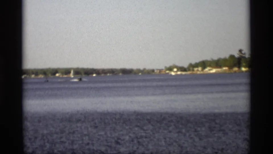 Home-movie frame of distant sailing activity on blue water near a wooded Holland, Michigan shoreline in 1969.
