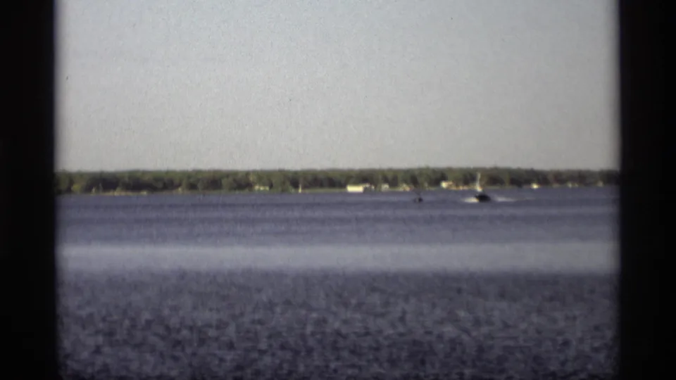 A steady 8mm lakeshore view shows small distant boats across the water, backed by trees and low structures in Holland, Michigan.