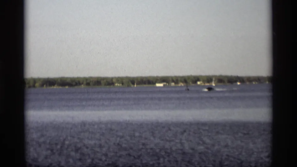 1969 Holland, Michigan 8mm frame of a small motorboat moving across open lake water near a tree-lined shore with scattered houses.