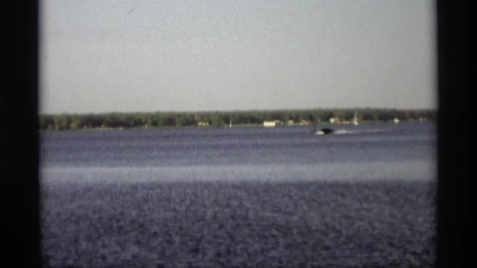 1969 Holland, Michigan lake view with a distant motorboat and white wake against a low wooded shoreline and small houses.
