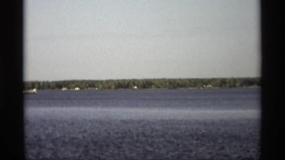 1969 Holland, Michigan home-movie frame showing rippled lake water, a wooded far shore, and small lakeside structures in the distance.