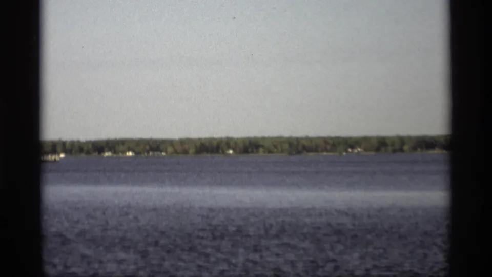 1969 Holland, Michigan 8mm view across open water toward a wooded shoreline with scattered houses and a dark frame edge.
