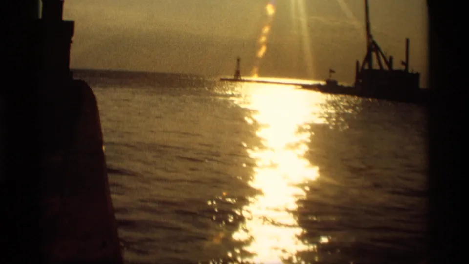 1969 Holland, Michigan 8mm frame showing golden sun glare across water and dark silhouetted structures near the horizon.