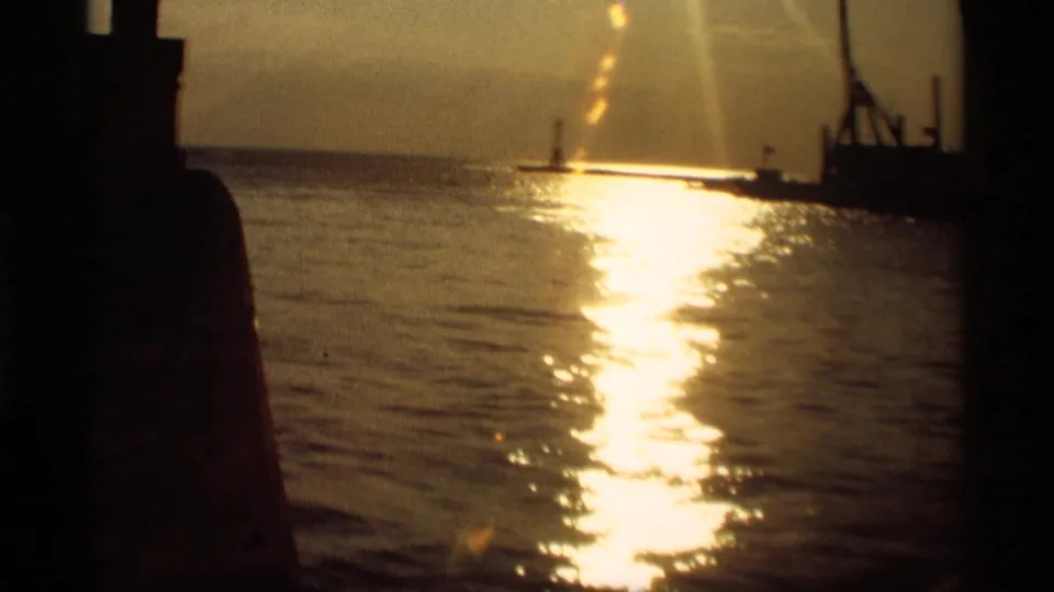 1969 Holland, Michigan home-movie frame with strong sunset glare on water and silhouetted waterfront forms.