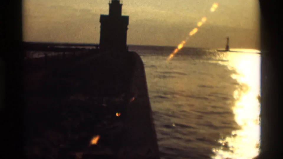 1969 Holland, Michigan 8mm frame showing a dark lighthouse silhouette, pier, bright water reflection, and distant beacon at sunset.