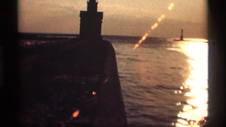 1969 Holland, Michigan lakeside view with the lighthouse in silhouette, a bright sun path on the water, and a small beacon offshore.