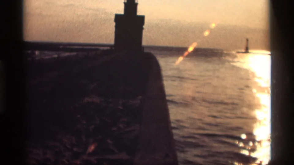 Vintage 8mm frame from Holland, Michigan showing a pier lighthouse in silhouette and strong sunset glare across moving lake water.