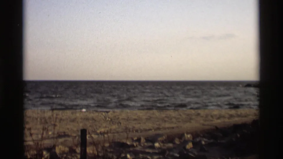 1969 Holland, Michigan shoreline view with lake horizon, pale sky, foreground grass, rocks, and dark 8mm frame edges.