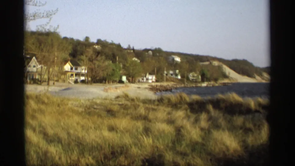 Home-movie frame of Holland, Michigan shoreline with grass, houses, trees, water, and a pale bluff or dune edge.