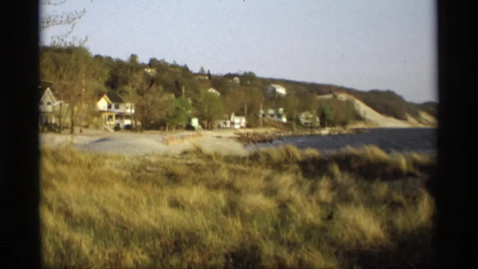 1969 8mm Holland, Michigan frame showing beach grass, lakeside houses, dark water, and a wooded hillside.
