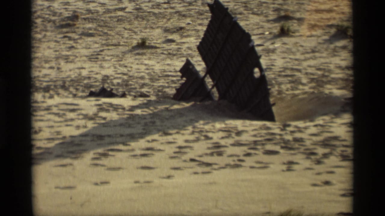 Weathered dark wooden structure half buried in sandy beach under warm light.