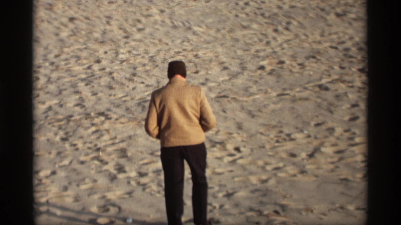 Man in a beige sweater walking away across a sandy beach.