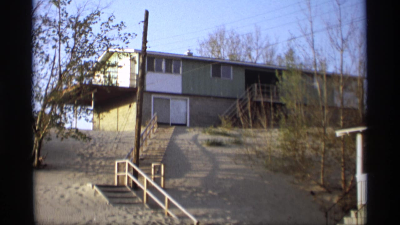 Tan and green split-level house above a sandy slope with white stair rail and trees.