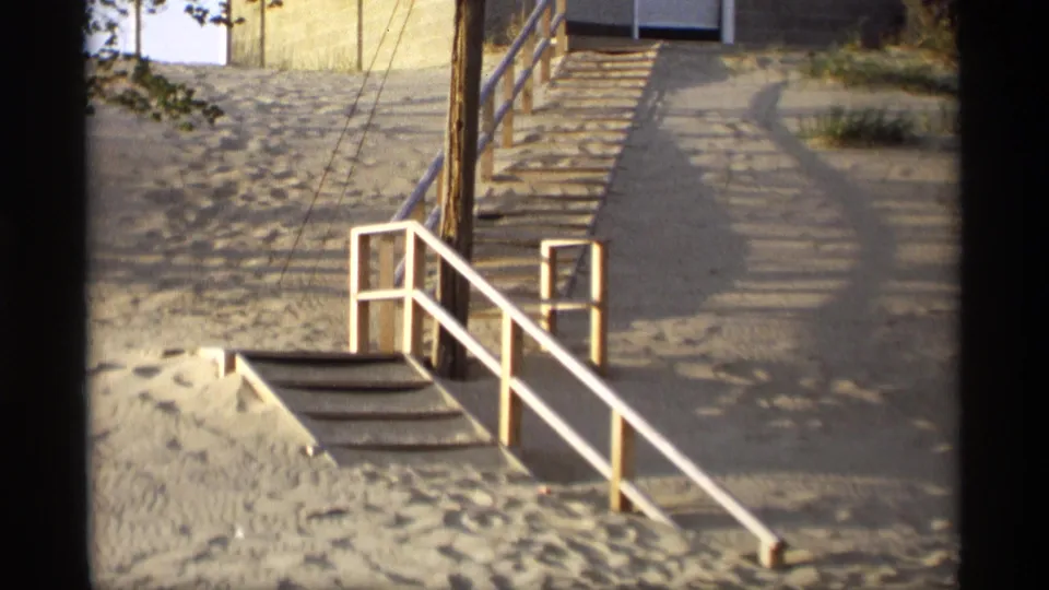 Closer 8mm frame of the stair approach below the Holland, Michigan house, showing white railings, wooden steps, sand, shadows, and a path.