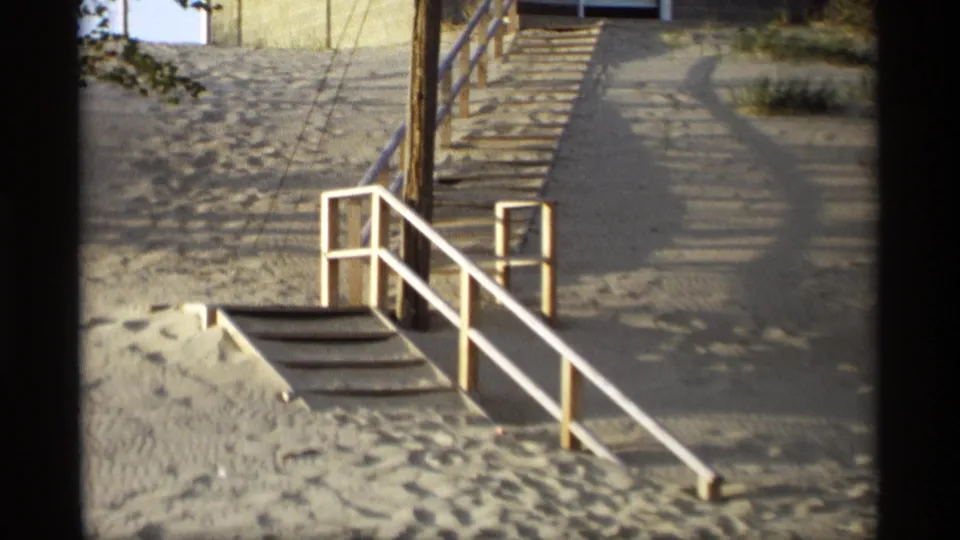 1969 Holland, Michigan home-movie frame showing a sandy beach access stairway with white railings, footprints, and a utility pole.