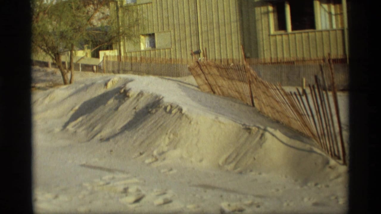 Sand dune beside a wooden fence and a building with vertical siding.